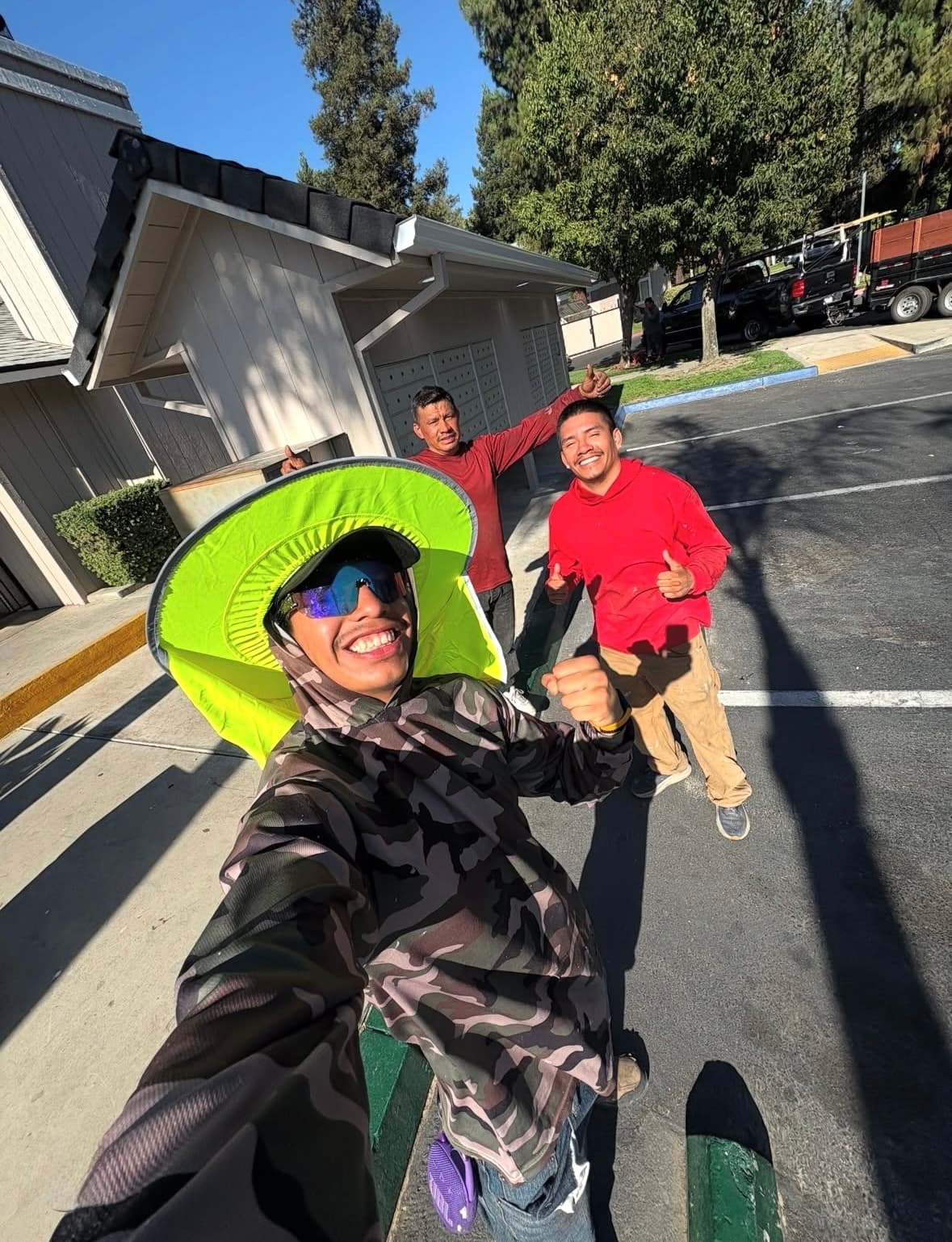 Smiling men, one in a bright green sun hat, pose for a sunny outdoor selfie.