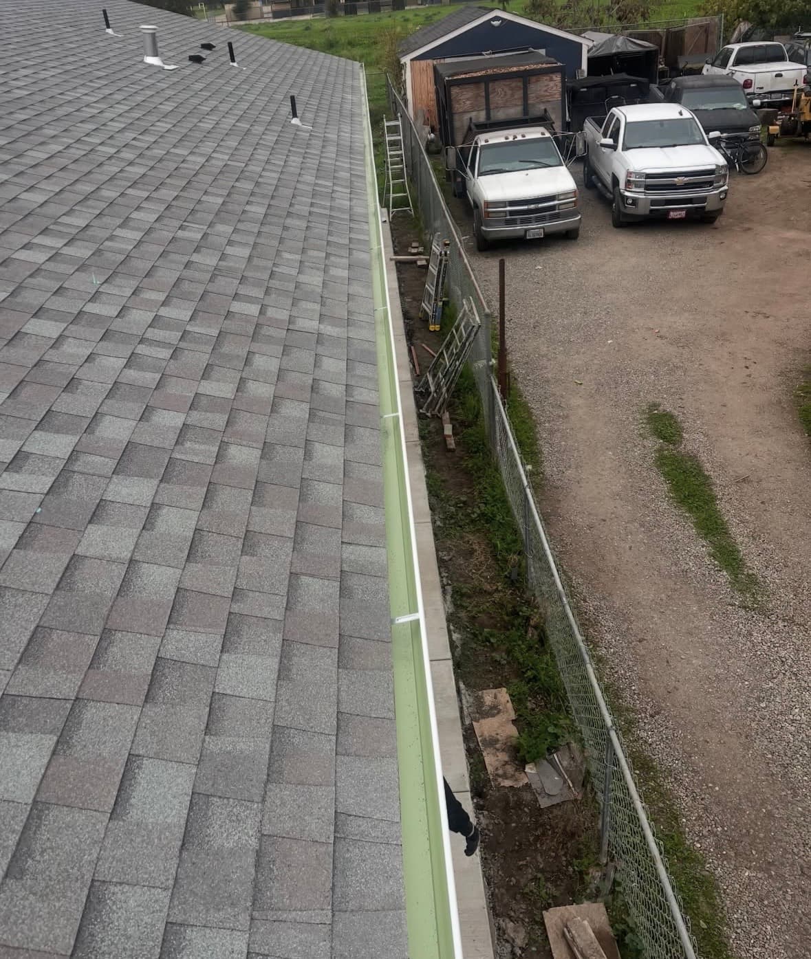 Gray shingled roof with light green gutter overlooking a gravel driveway with parked trucks.