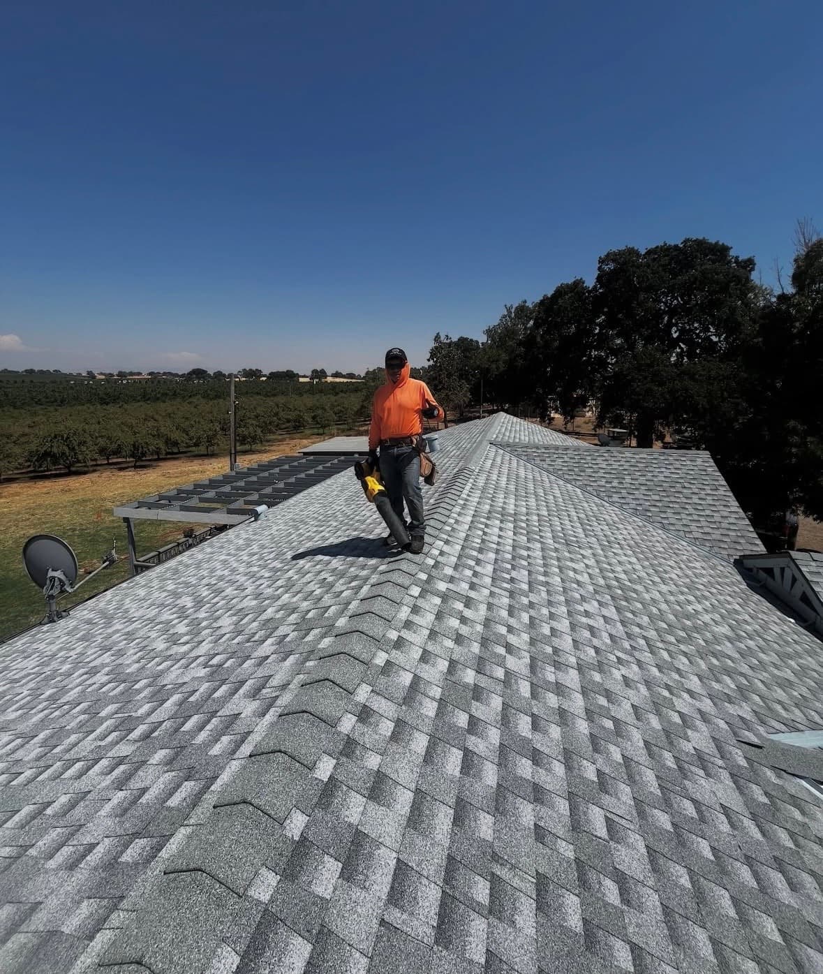 Roofer in orange hoodie on a new gray shingle roof under clear blue sky.