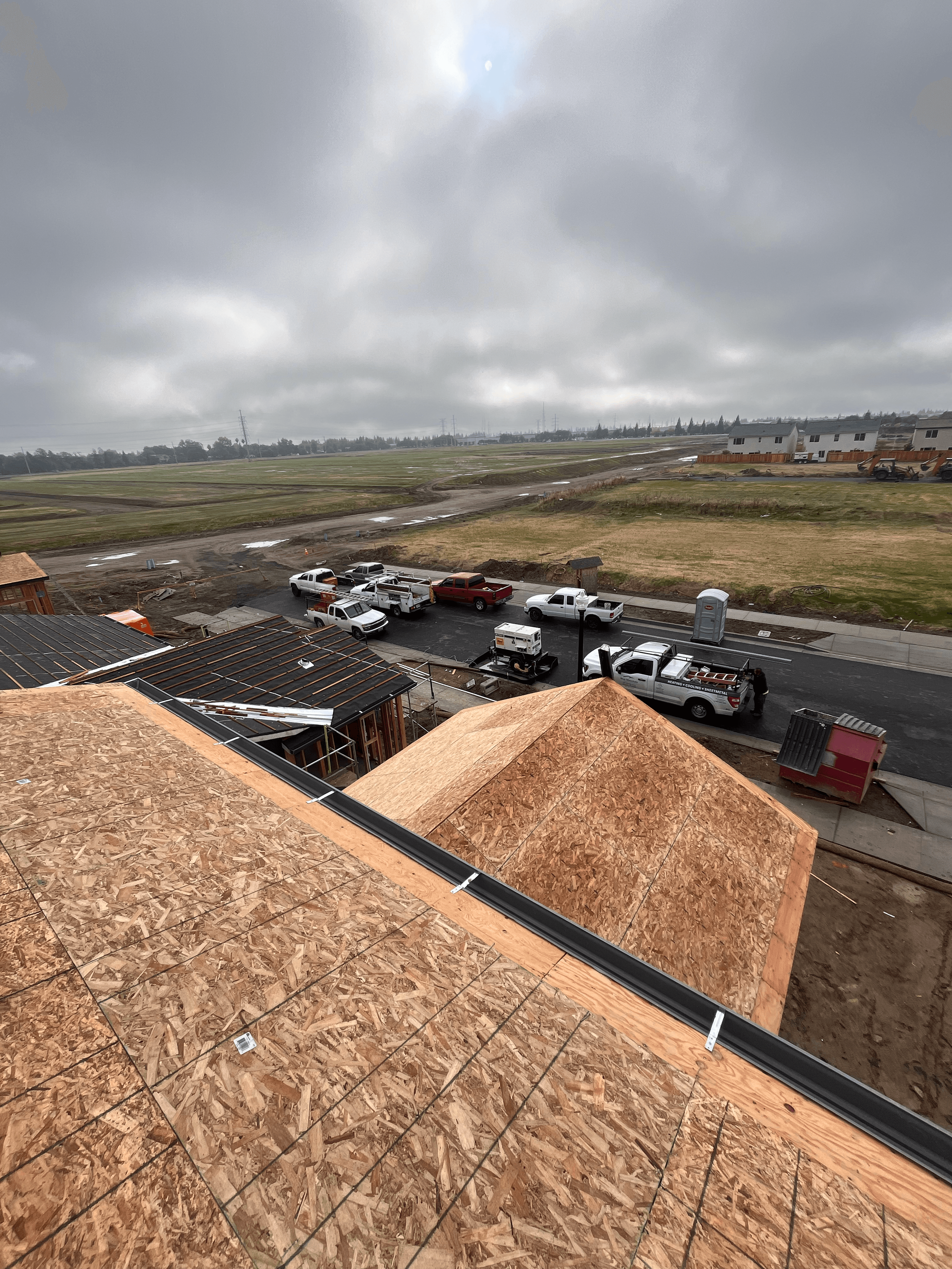 Rooftop view of new construction, work vehicles, and undeveloped land under a gray sky.