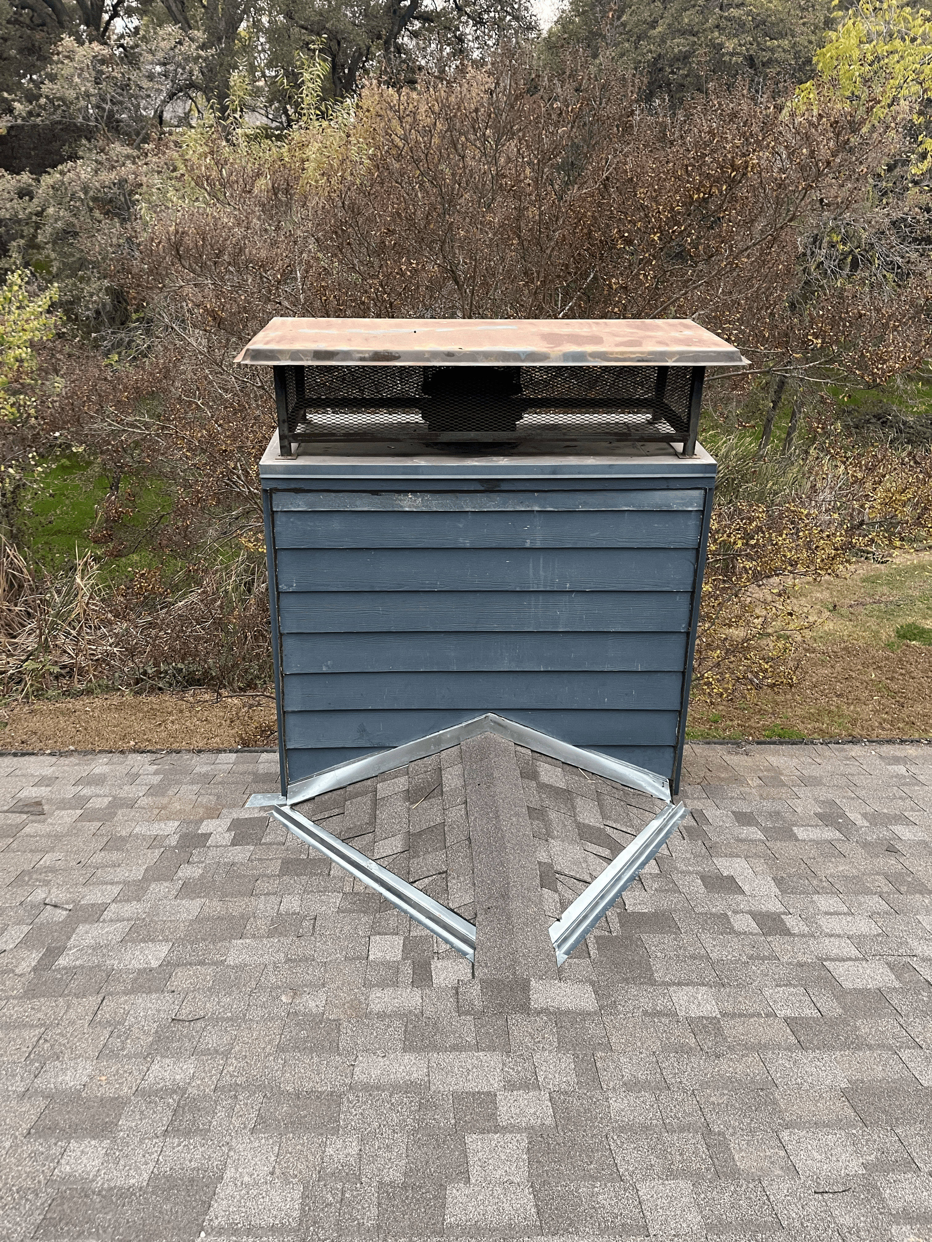Dark gray chimney chase with metal cap and flashing on a shingled roof, with trees.