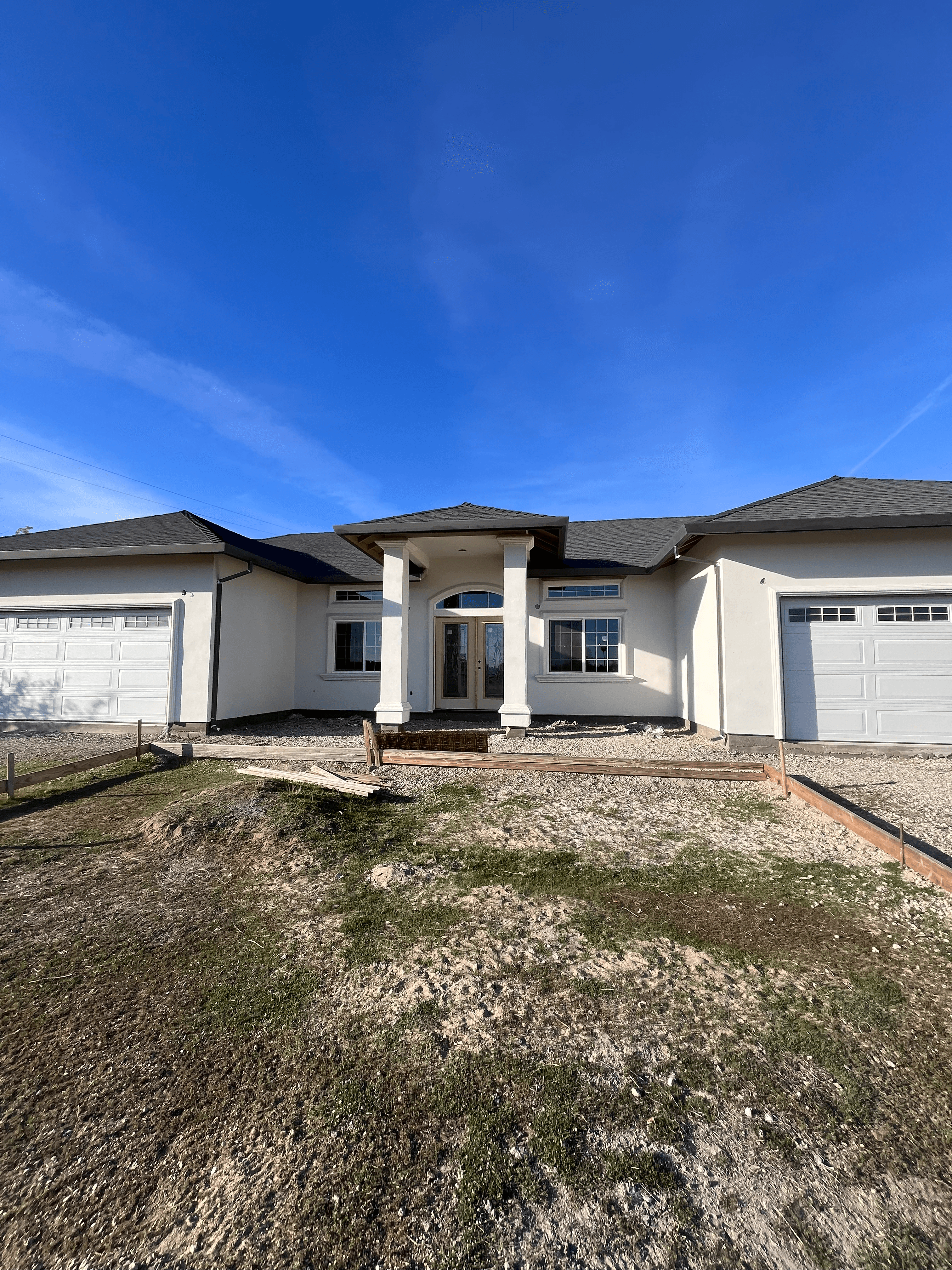 New white stucco house with columned entrance, two garages, and unfinished gravel yard.