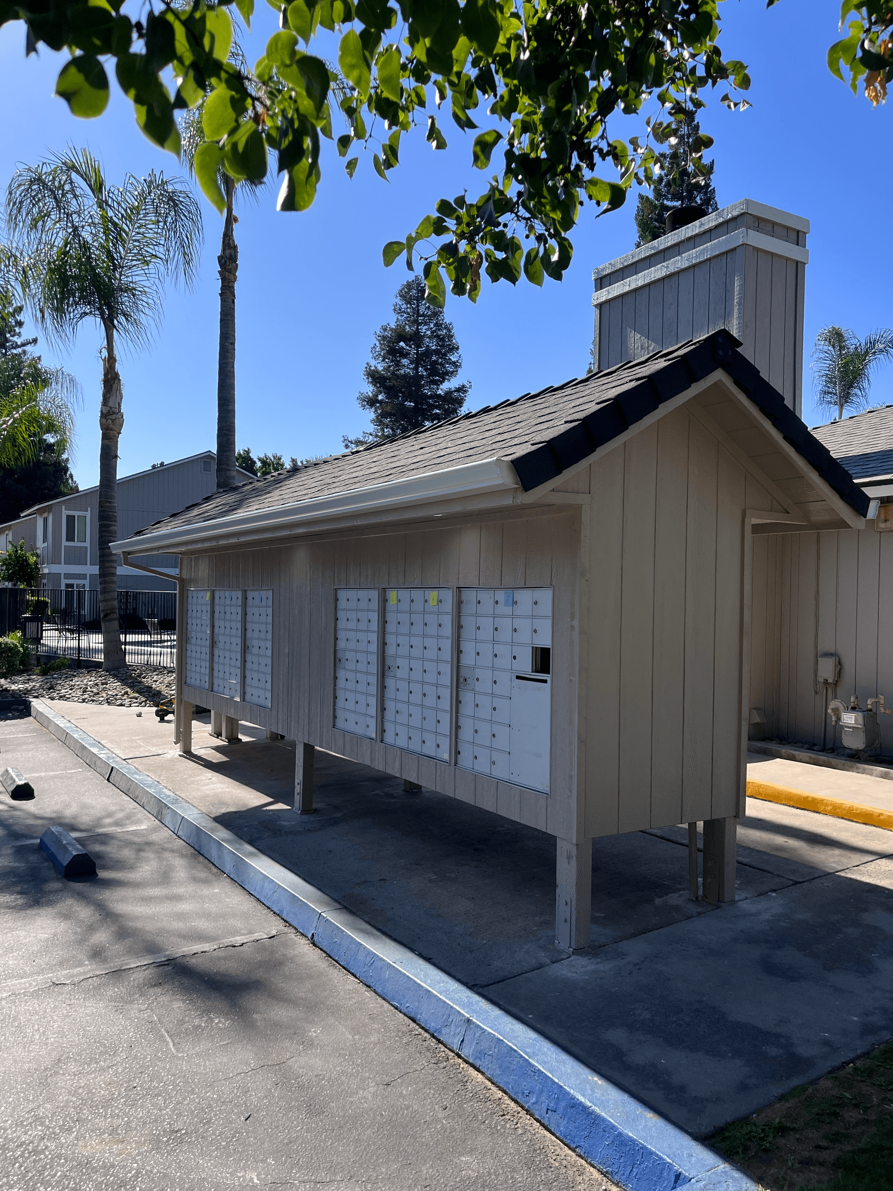 Light-colored community mailbox structure with many individual boxes, dark roof, blue sky, and trees.