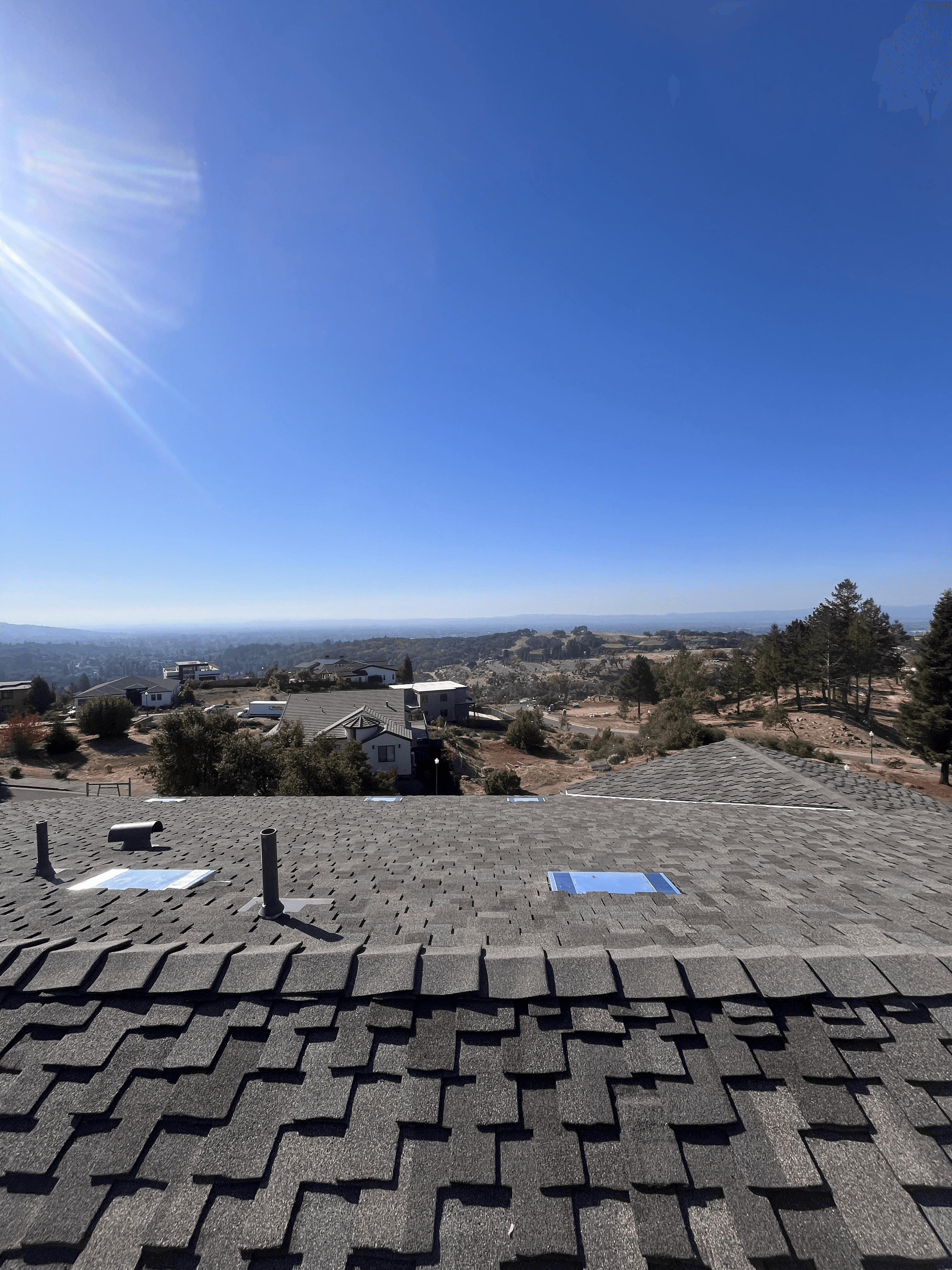Dark shingle roof with skylights, overlooking a valley of houses under blue sky.
