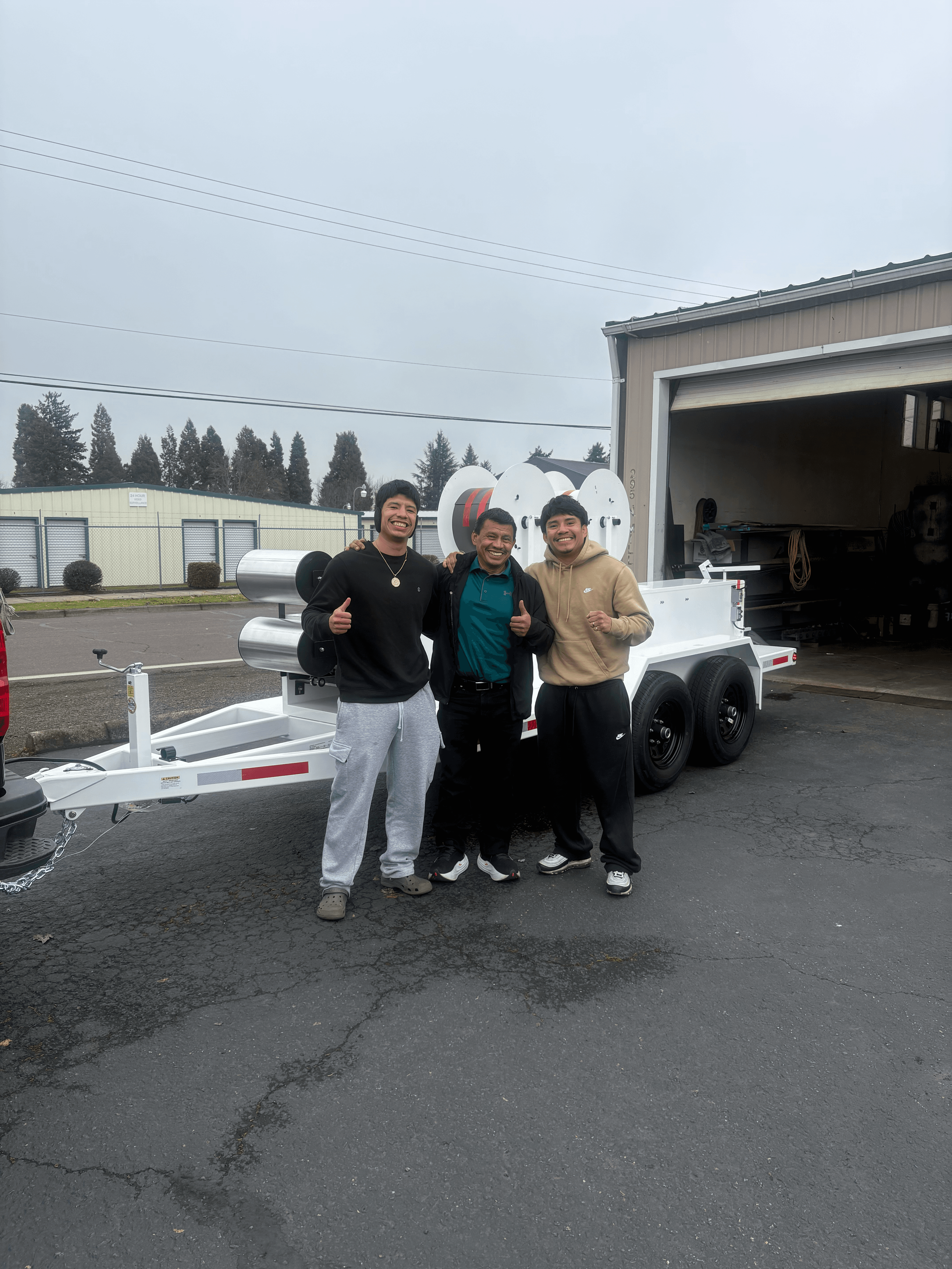 Three smiling men giving thumbs up next to a white trailer with large spools.