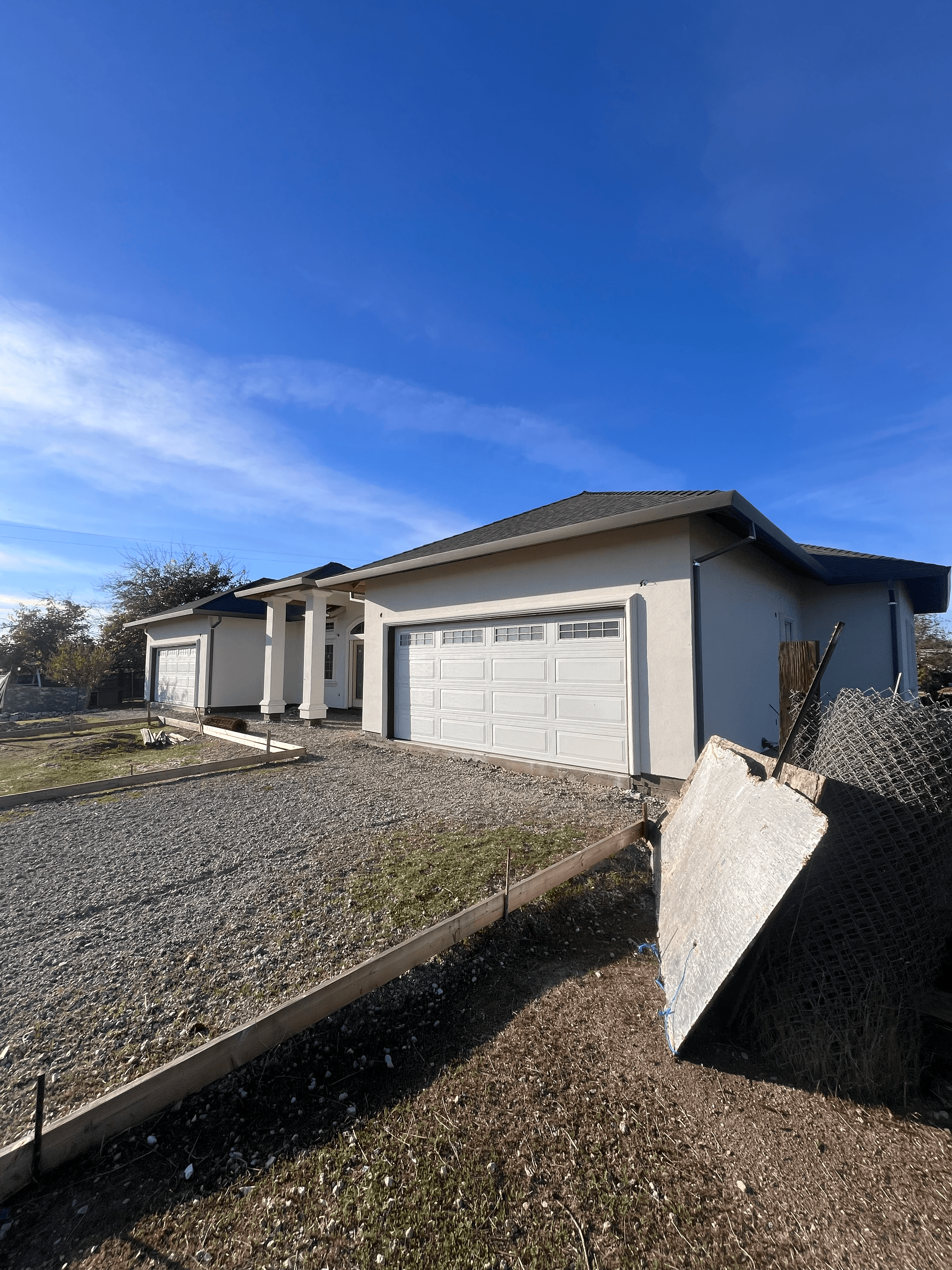 Light-colored house with white garage door, porch, gravel driveway, and fence under clear blue sky.