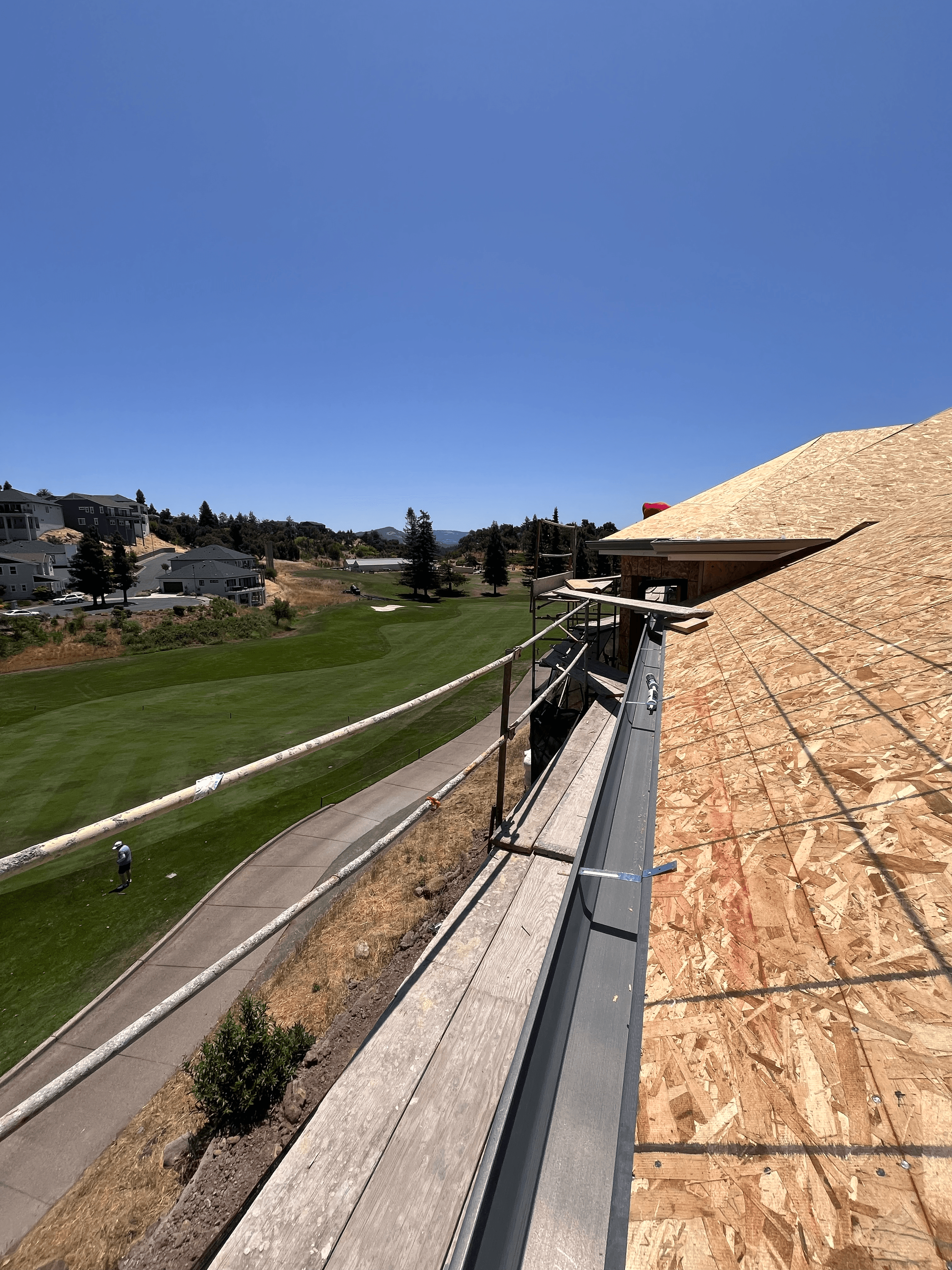 Scaffolding next to an OSB-sheathed building, overlooking a golf course and distant homes.